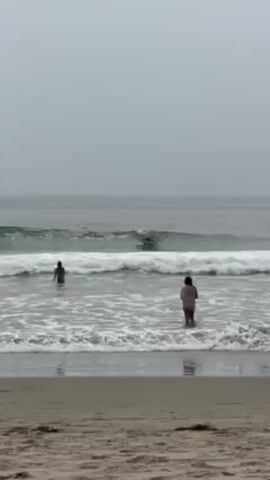 The video depicts a beach scene on an overcast day. The sea is grey with small waves breaking close to the shore. A surfer is riding a wave in the middle of the frame. Two people are standing in the shallow water, one person facing the surfer, and the other kneeling. The sand is visible in the foreground, leading up to the water's edge. The overall atmosphere is tranquil and somewhat muted due to the weather.