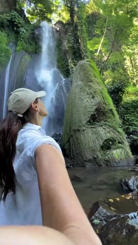 A woman with long dark hair, wearing a white shirt and a light-colored baseball cap, is taking a selfie in front of a waterfall. The waterfall cascades down a rocky, moss-covered cliff face surrounded by lush green vegetation. The water flows into a pool at the bottom, with rocks scattered around the edges. The woman is smiling and looking towards the camera.