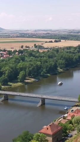 This is a vertically oriented aerial video of a river and a bridge. On the river, a white boat is visible. The bridge spans the river and connects two sides of land. On one side of the river, there are houses and trees. On the other side, there are fields and a distant town with rolling hills in the background. The sky is blue with a few clouds.