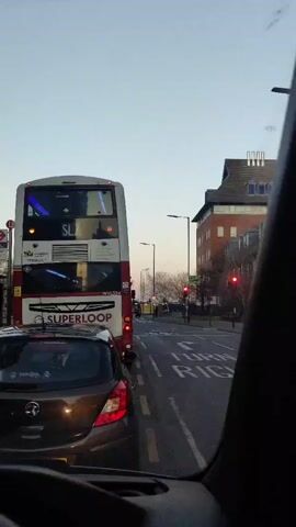 The scene is captured from inside a vehicle, with the steering wheel and dashboard visible. The view is forward-facing, looking out onto a street with stationary traffic. A double-decker Superloop bus occupies the lane ahead, with its rear facing the camera. Behind the bus, a dark-colored car is visible. The traffic lights are red. The street is marked with turn right signage. Buildings are seen in the background, and the sky is a clear blue with hints of sunset. The overall setting appears to be an urban environment, likely in the UK, as indicated by the signage and type of bus. No people are clearly visible.