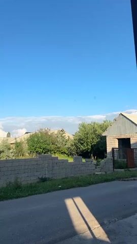 The video presents an exterior shot taken from a street, focusing on a residential area under a clear blue sky with a few clouds near the horizon. A low wall made of grey concrete blocks runs along the edge of the property, with varying degrees of completion. Green foliage, including trees and bushes, fills the space behind the wall, partially obscuring the roofs of houses in the distance. A small building, constructed from the same grey concrete blocks and topped with a corrugated metal roof, is visible to the right. Tall, slender trees rise in the background. Throughout the video, small birds flit across the scene. The road in the foreground is paved and displays a shadow cast by an unseen object.