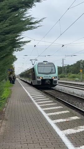 The video shows a train at a station. A train is moving from right to left on train tracks. The train is modern and has a light green and grey color scheme. The train has multiple cars and windows. On the left side of the screen is a platform made of interlocking bricks. Next to the platform is a hedge of green trees. Further down the platform, two people with luggage are standing. There are train tracks and overhead electrical lines above the train. The sky is overcast.