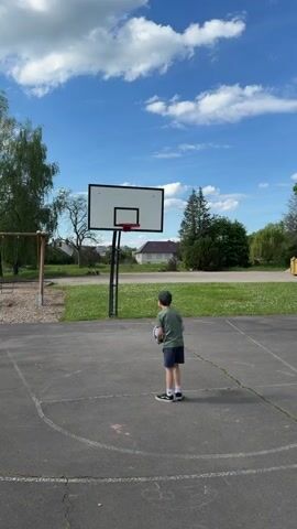 A young boy stands on a cracked asphalt basketball court, facing a basketball hoop with a white backboard and red rim. He wears a green t-shirt, dark shorts, and a baseball cap.  He throws the ball towards the hoop, which it misses. The sky is blue with puffy white clouds. A swing set is visible on the left, and grass and houses are in the background.