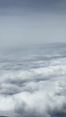 The video shows a view from an airplane flying above a thick layer of clouds. The clouds are white and fluffy, with some darker shadows visible, indicating depth and variation in their formation. The sky above the clouds is a light gray, suggesting an overcast or hazy atmosphere. The perspective is looking slightly downwards, giving the impression of vastness and isolation above the cloudscape. There are no people or discernible objects visible in the frame other than the clouds and the sky. The lighting is diffused and appears to be from daylight.