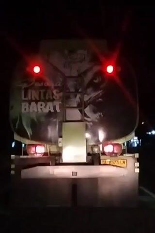 The video captures the rear view of a tanker truck at night, seemingly from a vehicle following it on a road. The scene is dark, with the truck's red taillights and white reverse lights clearly illuminated. The tanker itself features a large graphic design, prominently displaying the text "LINTAS BARAT" (meaning "West Crossing" or "Western Route" in Indonesian), along with what appears to be an eagle or bird of prey. The truck's license plate, "BH 8573 LN," is visible on the lower rear. Mudflaps are present beneath the bumper. On the left side of the frame, a yellow road sign with a black arrow pointing right is partially visible. The truck appears to be in motion, traveling along the road in the darkness.