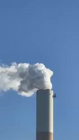 A vertical shot shows a tall smokestack releasing a plume of white smoke against a clear blue sky. The smokestack is predominantly white, with a beige-colored base, and has a small platform structure attached near its top. The smoke billows and disperses to the left side of the frame.