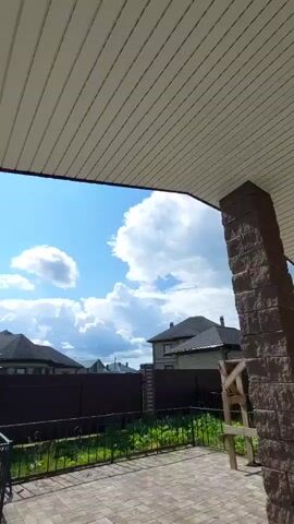 A video taken from a covered porch shows a view of a backyard and the sky. The porch ceiling is visible in the foreground, made of light-colored, ribbed material. To the right, a thick pillar constructed from dark, rough-textured stone supports the porch. The porch floor is paved with interlocking bricks in shades of brown and gray.

Beyond the porch, a low, dark brown metal fence encloses a grassy area with some greenery. In the background, residential houses with dark roofs are visible, separated by fences. The sky is a bright blue with scattered white cumulus clouds. The overall impression is of a sunny day in a suburban or rural setting. There are no people or animals visible in the video.