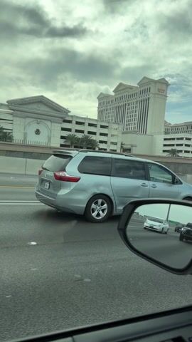The video shows a view from inside a car driving on a highway. On the left side, a large white building with the words "CAESARS PALACE" on top is visible. The building appears to be a hotel or casino. There's also a multi-story parking garage next to the building. In the side mirror of the car, other vehicles can be seen on the highway, including a white car and a dark gray Toyota Tacoma truck. The sky is cloudy.