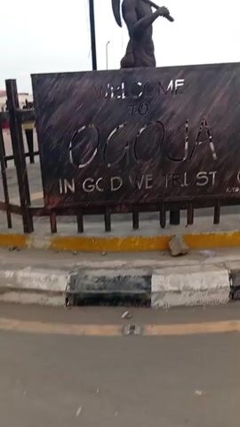 The video shows a sign welcoming people to "Ocoja" with the phrase "In God We Trust" written below. Atop the sign is a statue of a man wearing a hat and carrying a pickaxe over his shoulder. The sign is located behind a metal fence, which has red and white tape tied to it. The statue appears to be made of a dark metal or stone. The surrounding area seems to be a road or sidewalk. The weather is overcast.