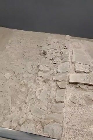 The video shows an archaeological site within a museum setting. The ground is covered in dirt and stone, with various remnants of ancient structures. In the foreground, a line of rough stones separates two distinct areas. To the left, the ground is more uniform and appears to be compacted earth. To the right, larger, flat stones are laid out, some appearing to be broken or incomplete. As the camera pans and moves, the remains of walls built with layered stones and what appears to be plaster are visible. There are also stone columns and sections of flooring. The lighting suggests an indoor exhibition space, with track lighting illuminating the artifacts. No people are visible in the video.