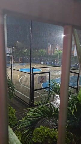 The video shows an outdoor basketball court during a heavy rainstorm at night. The court is surrounded by a fence with a sign that reads "FAVOR MANTENER LA REJA CERRADA" (Please keep the gate closed). The court is lit by overhead streetlights, which reflect brightly off the wet surface. Behind the court, there is a row of parked motorcycles and a building in the distance. The camera is positioned behind a metal railing and some greenery, partially obscuring the view. The heavy rain is visible in the foreground and across the entire scene.