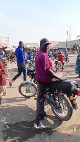 The video shows a busy street scene in what appears to be a market area. Several people are visible, including men and children, with some standing near motorcycles. The man in a purple shirt and cap is sitting on a motorcycle, looking towards the camera. Other men are riding motorcycles through the scene. Market stalls with umbrellas are visible in the background, suggesting a commercial activity. The ground is paved, with some debris and shadows visible. Overall, the video captures a typical day in a bustling market environment.