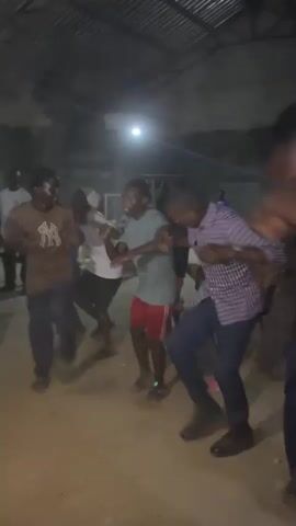 This media file captures a lively gathering of people dancing indoors. The scene appears to be a rustic, dimly lit space with a corrugated metal roof. The floor is dirt or sand, suggesting an outdoor structure or a simple indoor space. A single bright light source illuminates the center of the activity, casting shadows and creating a dynamic atmosphere.

Several individuals are visible, mostly men, engaged in energetic dancing. They are dressed in casual attire. One person in the foreground is wearing a light brown t-shirt with a white graphic. Another dancer wears a light blue t-shirt and red shorts. A person in a checkered shirt and dark pants is also visible. The focus is on the movement and the collective energy of the dancers.

The time of day is likely evening or night due to the artificial lighting and the overall dimness of the surroundings. There are no clear indications of the weather. The setting in Gudi, Nigeria, provides a cultural context for the gathering, suggesting a community event or celebration.

The emotions conveyed are primarily joy and exuberance, evident in the dynamic poses and apparent movement of the dancers. The blurry nature of some of the figures emphasizes the fast-paced action. There is no visible text within the media file. The overall impression is one of vibrant community spirit and enjoyment.