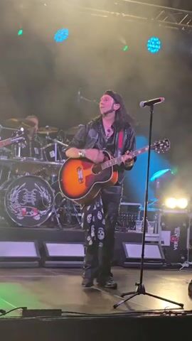 The video shows a live music performance on a stage illuminated by blue and green lights. A male guitarist with long dark hair and wearing a black shirt, vest, and black pants with skull patterns is playing an acoustic guitar and smiling. He is standing center stage. Behind him, a drummer is visible, and part of a drum kit with a logo is seen. Other band members and equipment are also present on stage. The lighting changes throughout the clip, with blue lights dominating at times. In the final few seconds, another person wearing a cowboy hat briefly appears on the left side of the frame. The overall atmosphere appears energetic and celebratory.