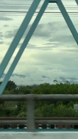 The video shows a view from a moving vehicle, likely a car, as it crosses a bridge. The perspective is through a light blue metal structure that forms part of the bridge's railing or support. The background reveals a landscape with lush green trees, rolling hills, and a cloudy sky. In the distance, mountains are visible, partially obscured by mist or clouds. The vehicle is moving from left to right, and the passing scenery is slightly blurred due to motion. A single tall tree stands prominently in the middle ground. Towards the end of the clip, a small, light yellow building with a dark window and a red-tiled roof comes into view on the right side of the frame, also blurred by the movement. The overall impression is one of a scenic journey through a rural or natural environment.