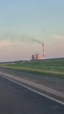 The video shows a view from a moving vehicle, looking out at a large industrial complex with a tall, striped smokestack emitting smoke against a clear sky. The complex includes a large, round cooling tower. The vehicle is traveling on a paved road, with a green grassy field to the right of the road. A triangular road sign with a figure on it is visible in the field. The lighting suggests it is either early morning or late afternoon.
