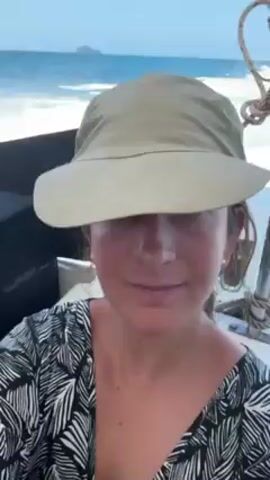 A woman on a boat, wearing a tan hat and a black and white patterned shirt, is looking into the camera. The boat is moving through the water, creating waves behind it. In the distance, a small island can be seen. There is rope to the right. The sky is blue.