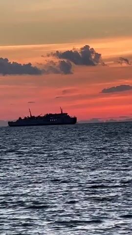 A wide shot shows a ferry boat on the ocean under a dramatic sunset sky. The sky is a gradient of orange, pink, and red, with scattered dark clouds. The ferry is a dark silhouette against the vibrant sky. The ocean is dark blue with choppy waves. The ferry appears to be moving slowly or is stationary. There are no people visible on the ferry or in the water. The scene is tranquil and atmospheric.