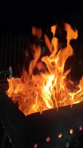 The video shows a close-up view of a bonfire in a metal grill. The fire is intense and produces tall, flickering flames in shades of orange and yellow against a dark background. Some logs are visible within the grill, partially consumed by the flames. The grill itself is dark and appears to be made of metal, with circular holes along its base. The scene is dark, suggesting it is nighttime or a dimly lit environment. No people are visible in the frame.