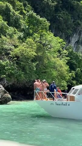 A group of people are standing on the bow of a white boat. The boat is in turquoise water with a lush green cliff face in the background. The people are wearing swimwear and appear to be on a tour. One person is holding a phone, possibly taking a picture or video. There are about 5-7 people on the bow, and at least two crew members are visible on the boat. The boat has Thai writing on its side.