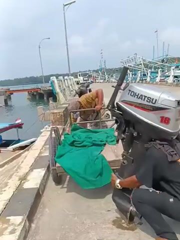 Two men are on a concrete dock near the water. One man, wearing a brown shirt and a hat, is leaning over a small cart with a green tarp on it. The second man, wearing a black t-shirt and black pants, is handling a large outboard motor. The motor has the brand name "TOHATSU" and the number "18" visible on it. In the background, there are boats docked, a pier with railings, and some trees and buildings on the distant shore. The sky is bright and overcast. The man in the black shirt picks up the outboard motor and places it on his shoulder.