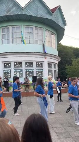 A group of young people, all wearing blue t-shirts and light-colored pants, are performing a dance routine outdoors in front of a building. The building is light blue with white trim and has a distinctive green, pointed roof. The dancers are arranged in rows, with some in the foreground and others further back on a raised platform or stage. In the background, there are windows on the building, and a Ukrainian flag is visible. There are speakers and what appears to be DJ equipment on the stage. The ground is paved with gray stones. The overall atmosphere appears to be that of an outdoor performance or event.