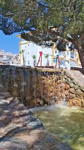 A small, tiered waterfall cascades over rocks into a pool of water. The water appears clear and slightly rippled. Lush green foliage, likely from a large tree, overhangs the scene from the top left. In the background, there is a bright white and yellow building with multiple windows and balconies. Adjacent to the building, a red and yellow swing set is visible. Several palm trees stand between the waterfall and the building. The setting appears to be an outdoor public space, possibly a park or a courtyard, given the presence of the waterfall and playground equipment. The sun is shining brightly, indicating it is daytime, likely late morning or early afternoon, and the weather is fair. The scene evokes a sense of tranquility and relaxation due to the gentle sound and visual of the flowing water, contrasted with the cheerful appearance of the playground. No people are visible in the frame. There is a white sign with a green symbol on it on the left side of the frame, near the palm trees.