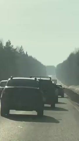 The video shows a line of cars driving down a highway, flanked by a forest on either side, on a bright, sunny day. The camera is presumably inside one of the cars, positioned behind the first few vehicles. The cars, mostly SUVs, are spaced out but moving in a convoy-like formation. The road appears to be relatively straight with a slight curve in the distance. The camera shifts abruptly and there's heavy water splash on the windshield, obscuring visibility.