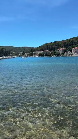The video shows a clear blue sky over a body of water. The water is crystal clear, revealing a rocky seabed. In the distance, along the shoreline, there are houses and trees. Several sailboats are visible on the water. The overall scene depicts a sunny day in a coastal area.