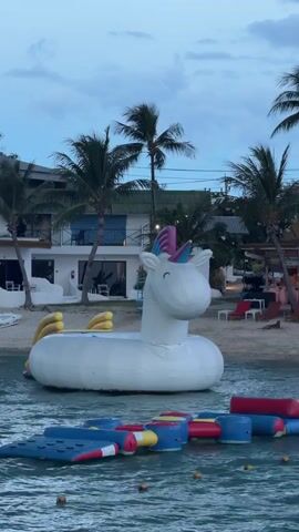 A large, inflatable unicorn pool float is situated in the shallow waters of a beach. Behind the unicorn, a beach with palm trees and white buildings with balconies is visible. In the foreground, a colorful inflatable water park structure is floating in the water. The sky is overcast.