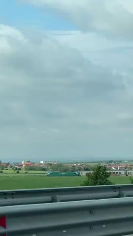 The video, shot from inside a car, shows a green and white train moving through a rural landscape. The train, passing to the left, is adjacent to a green field. A small village can be seen in the distance, under a cloudy sky with patches of blue. The car is moving along a highway, with a gray guardrail visible in the foreground. The camera is static and the shot is focused on the train and surrounding landscape.