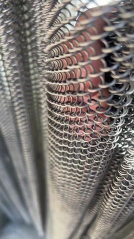 A close-up shot shows a person's fingers visible through a dense chainmail curtain or fabric. The chainmail is made of small, interlocking metal rings, creating a woven texture. The fingers are slightly blurred due to the focus on the metal mesh. The lighting suggests an indoor environment, possibly with natural light filtering through the chainmail. The camera angle is low, looking upwards at the chainmail. The overall effect is that the fingers are partially obscured and distorted by the chainmail texture.