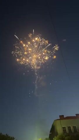 A static low-angle shot captures fireworks exploding against the dark blue sky, partially obscured by a house's rooftop on the right. The fireworks are in shades of red, white, and gold, bursting into patterns. A bright, green-yellow orb glows beneath the fireworks, emitting a trail of smoke. Two thin wires run diagonally across the top right corner. The house, with a reddish-brown tiled roof and cream-colored walls, is visible at the bottom right, with visible windows.