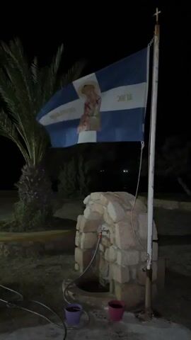The video opens with a medium shot of a flag flying on a flagpole at night. The flag is blue and white with a depiction of a religious figure, possibly a saint, and some text that is difficult to read in the low light. The flag is attached to a white pole topped with a cross. In the foreground, there is a small, rustic stone fountain with a spigot and a hose attached, leading into a basin. Two small potted plants, one purple and one pink, are placed near the basin. To the left of the fountain, a palm tree with large fronds is visible. The background is dark, with some blurry lights in the distance, suggesting an outdoor setting at night. The flag billows gently in the wind. The video is static for the first few seconds, then there is a slight shift in perspective and the flag continues to wave. There are no people visible in the video.