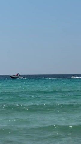 The video shows a bright, sunny day at sea. The water is a vibrant turquoise near the shore, transitioning to a deep blue further out. Small waves are visible. Two boats are in motion. The first, a white speed boat, is closer to the shore and appears to be towing something or someone. It has several people on board, all wearing orange life vests. The second boat, further out and smaller, is difficult to make out clearly but seems to be a personal watercraft. The sky is clear and blue.