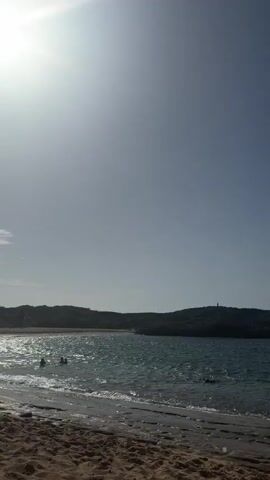 The video shows a beach scene on a sunny day. The camera is positioned on the sand, looking out towards the ocean. In the foreground, there is a sandy beach with some rocks at the water's edge. The water is blue and choppy, with small waves breaking on the shore. In the background, there are hills covered in vegetation, and a clear blue sky. Several people are visible in the water and on the beach. Two men are standing in the water, facing away from the camera. Further out in the water, there are other people swimming and wading. On the beach, two people are sitting down, facing the water. The sun is bright and high in the sky, creating a lens flare in the upper left corner of the video. The overall impression is one of a peaceful and relaxing day at the beach.