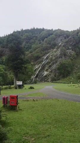 The video shows a scenic view of a waterfall cascading down a rocky mountainside. The waterfall is surrounded by lush green trees and vegetation. The foreground features a well-maintained grassy area with a paved path winding through it. Two red mailboxes are visible on the left side of the path, and a signpost stands nearby. The sky appears overcast, suggesting a cloudy day. The camera pans slightly upwards, focusing on the waterfall and the mountainous terrain.