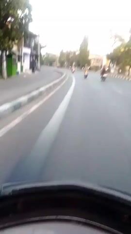 The video shows a first-person perspective of someone riding a motorcycle on a paved road. The road appears to be in a semi-urban or suburban area, with buildings and trees lining the sides. There are several other motorcycles visible on the road ahead, also moving in the same direction. The rider's dashboard is visible at the bottom of the frame, showing a speedometer with a needle indicating a speed of around 40-50 units. The time stamp indicates the video begins at 00:00 and ends at 00:05. The lighting suggests it is daytime, possibly late afternoon due to the angle of the sunlight.
