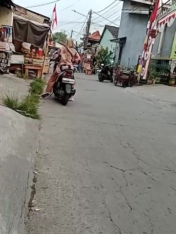 A person on a scooter is facing away from the camera on a street. They are wearing a headscarf and a long-sleeved shirt and pants. There are shops and houses on either side of the street. There are other people and vehicles in the background. The street is made of asphalt and has some cracks in it. There is a sidewalk on the left side of the street. The sky is overcast.
