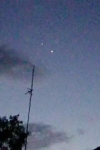 The video displays a dim, twilight sky with two bright, luminous objects. A tall, thin, dark antenna stands in the foreground, bisecting the frame vertically and extending towards the bottom edge. A portion of a dark tree is visible in the lower left corner. The sky is a gradient of blues and grays, with some wispy clouds. The two celestial objects, one slightly higher and to the left of the other, appear to be planets or stars. The object on the right glows with a yellowish-white light, while the object on the left appears slightly dimmer and whiter. There are no people or visible interactions in the video. The scene is static with no discernible movement apart from the subtle shifting of the camera, causing a slight blur.