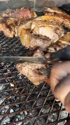 The media displays a close-up shot of various cuts of meat, primarily beef, cooking on a heavy metal grill grate over glowing charcoal. The background reveals a rustic brick wall, suggesting an outdoor or semi-enclosed grilling area, typical of an Argentine "asado."

In the foreground, a dark-skinned hand uses a carving knife with a wooden handle and a metal fork to cut into a piece of meat directly on the grill. The cut reveals a reddish-pink interior, indicating a medium-rare to medium doneness, with the exterior of the meat showing a well-browned and slightly charred surface. Other pieces of meat, including what appear to be ribs and thick steaks, are also visible on the grill, some appearing more cooked than others. Rendered fat glistens on the meat.

The activity is the preparation and sampling of grilled meat, a central part of an asado, consistent with the cultural context of Merlo, Argentina. The bright lighting suggests it is daytime or a well-lit environment. There is no visible text in the image.