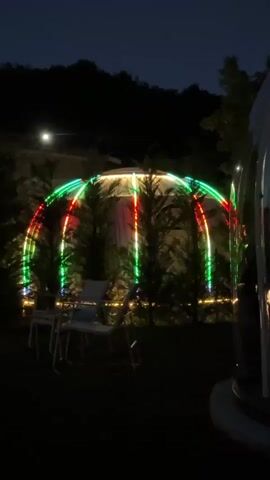 The video shows an outdoor scene at night. In the center, there is a large geodesic dome structure illuminated with colorful LED lights in red, green, and white. The lights appear to be arranged in vertical stripes and curved bands, creating a festive atmosphere. Several evergreen trees are planted in front of the dome, their dark silhouettes contrasting with the bright lights. Two white chairs and a small table are placed in the foreground, suggesting a seating area. The background features a dark hillside or mountain, sparsely populated with trees, and a distant light source, possibly a streetlamp or house light. The overall impression is that of a decorated outdoor space, perhaps a campsite or backyard, set up for an evening gathering.