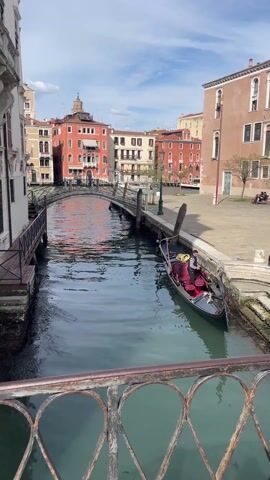 The video shows a scenic view of a canal in Venice, Italy. The canal is narrow, and the water is slightly murky. On the right side of the canal, a black gondola with red interior is docked near a stone embankment. A gondolier in a striped shirt and dark pants stands on the embankment next to the gondola. Behind the gondola is a small square with some people sitting on benches.  Across the canal, a narrow, arched footbridge spans the waterway. The buildings surrounding the canal are varied in color and architecture, primarily in shades of red, yellow, and white. The sky is blue with some scattered clouds. The overall lighting is bright and sunny.