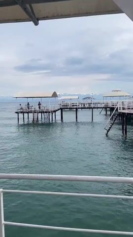 The media shows a view from a boat approaching a wooden pier on a cloudy day. The pier extends into the turquoise water, supported by numerous wooden posts. Several covered seating areas with white railings are visible on the pier, suggesting it is a recreational area or a place for boats to dock. Some people are seen on the pier, appearing relaxed. The sky is filled with dramatic, grey clouds, hinting at potential rain or a storm, but a sliver of brighter sky is visible on the horizon. The overall atmosphere is serene, with the gentle ripples on the water contributing to a peaceful mood. The location is identified as Choktal, Kyrgyzstan. There are no discernible text elements within the visible portions of the media.
