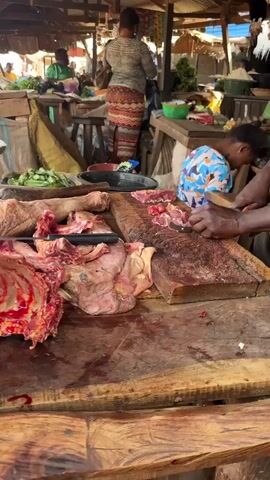 This is a medium shot of a butcher cutting meat at a wooden stall in an outdoor market. The butcher, a man with dark skin wearing a brown shirt, is using a cleaver on a large piece of meat. The meat is bloody and partially cut, revealing bones and muscle. In the background, other people can be seen at market stalls, and there are piles of vegetables and other goods. The environment appears to be a busy, open-air market.