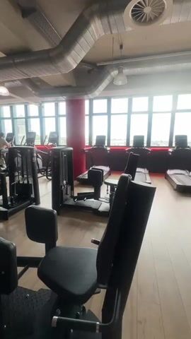 The video shows a gym with rows of exercise machines. In the foreground, there are two black weight machines with padded seats and backrests. One of the seats is pink. In the background, there are treadmills, and other weight machines, some with red accents. The gym has large windows letting in natural light. Exposed pipes and ventilation systems are visible on the ceiling. One person, wearing dark clothing, is visible in the distance, standing near a weight machine. The floor is wooden.