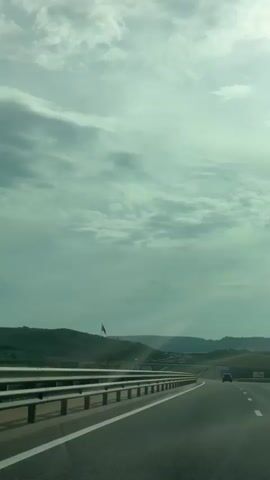 The video shows a vehicle driving on a highway. The sky is cloudy, and the surrounding landscape is green and hilly. There is a highway sign on the right side of the road, and a flag visible in the distance on the left side. Other vehicles can be seen on the road in the distance. The vehicle continues to travel along the highway throughout the video.