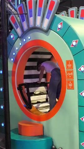 A young female child is climbing up a sloped, padded staircase inside a colorful, amusement park-like structure. The structure is predominantly teal and orange, with red and blue decorative elements at the top and blue lights around the circular opening. The staircase has black and white striped padding. A black handrail is visible to the left of the child. To the right of the opening, there are signs with text in Russian indicating age and height restrictions. The child is wearing a white t-shirt and dark pants. The environment appears to be an indoor play area.