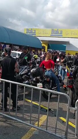 The video depicts a lively gathering of people and motorcycles in a parking lot next to a "Daka Se Feliz" store. The environment is outdoors, with partly cloudy skies overhead. A variety of motorcycles are parked throughout the area, with individuals standing and interacting amongst them. Some people are wearing motorcycle gear, while others are in casual attire. One woman in a red shirt is sitting on a motorcycle. There is a building that resembles a steakhouse in the background. People are socializing, and the atmosphere appears festive. The scene captures a motorcycle meetup or rally.