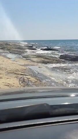 The video shows a view from inside a car looking out at a rocky shoreline. The car appears to be driving slowly over shallow water that washes over the rocks. The ocean is visible in the background, with waves breaking on the shore. The sky is clear and blue. A Mercedes-Benz emblem is visible on the hood of the car.
